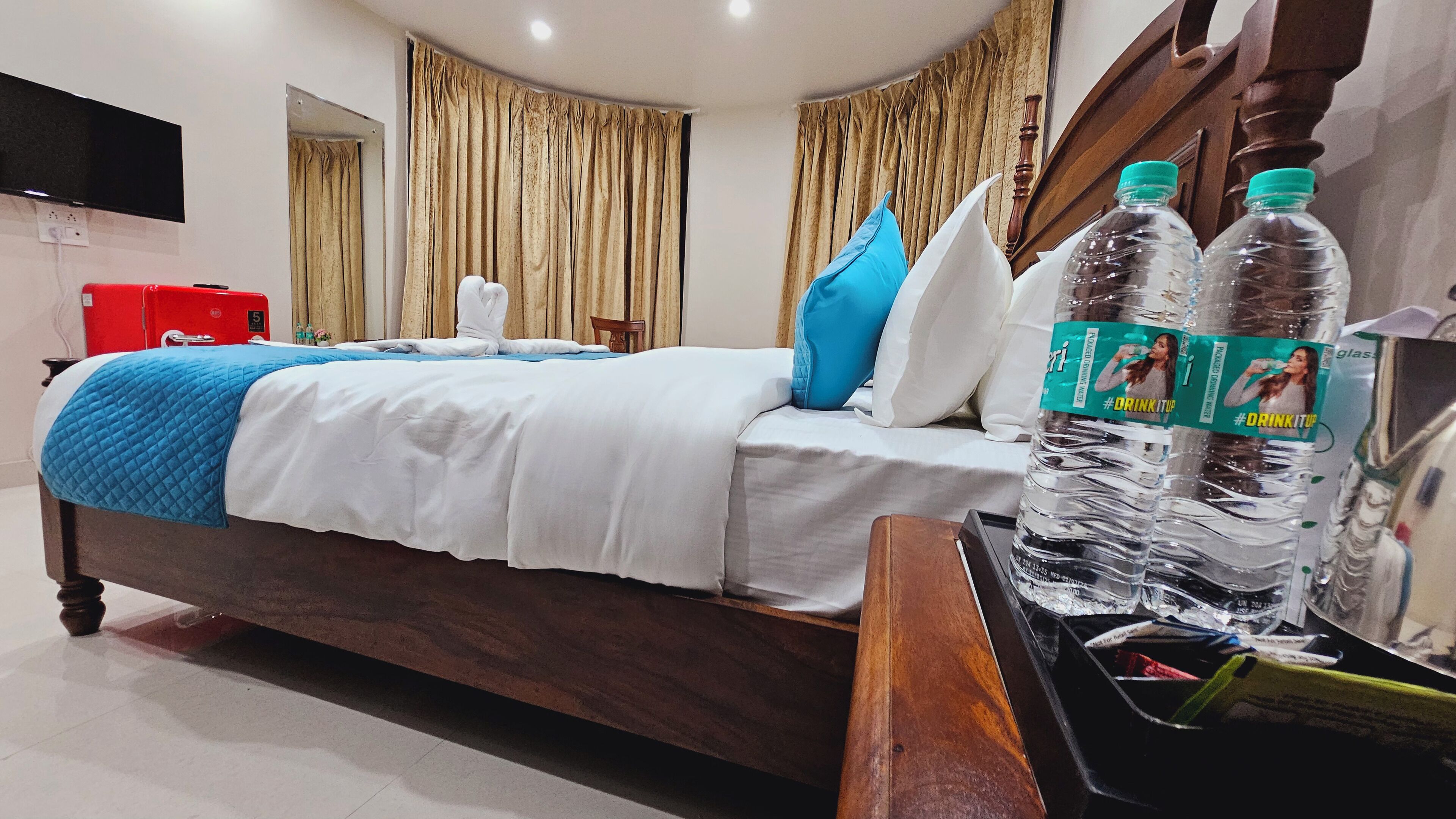A hotel room with a bed, a red suitcase, and two water bottles on a table.