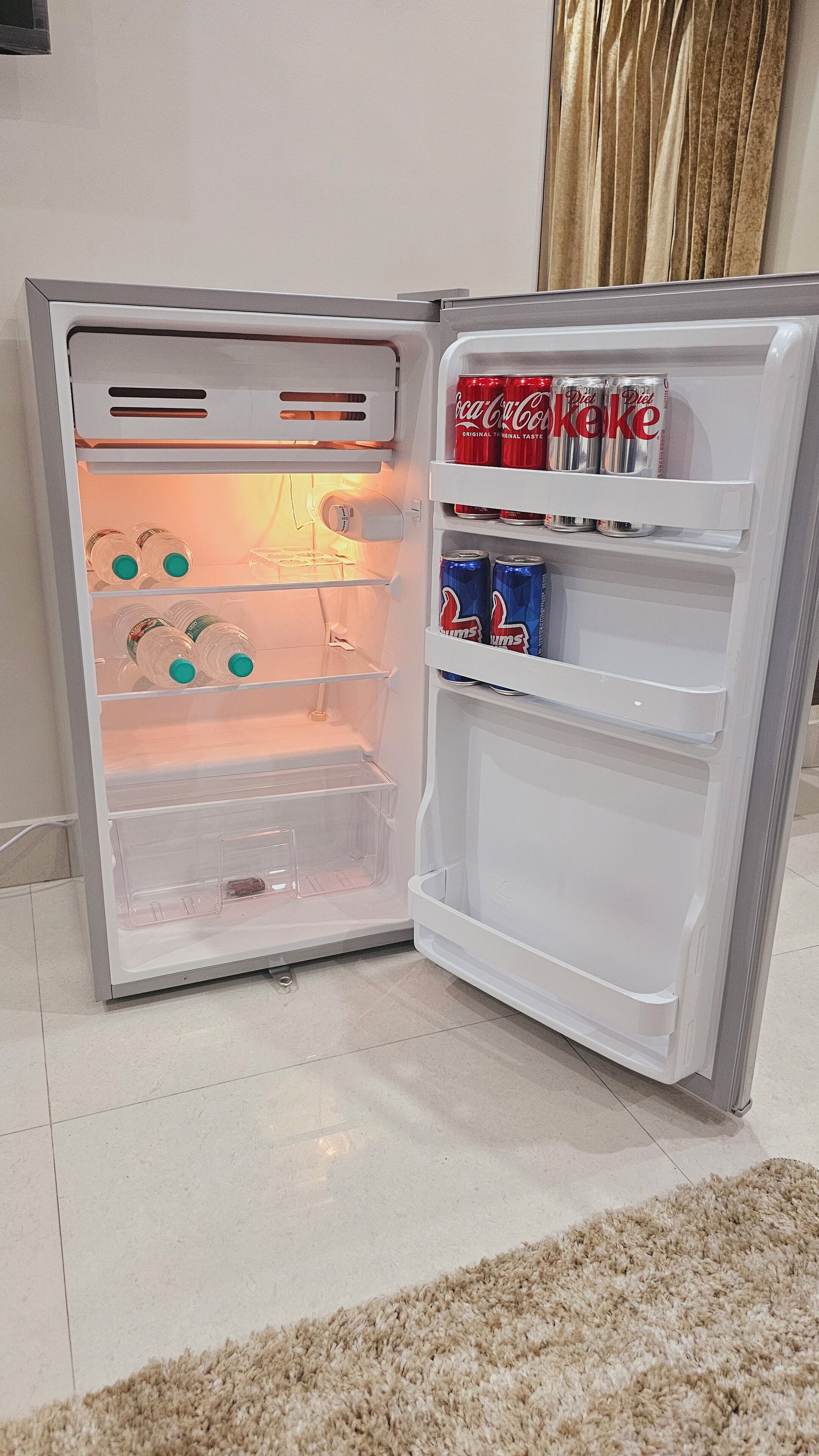 Un réfrigérateur ouvert, avec des étagères contenant des boissons et une bouteille d’eau.