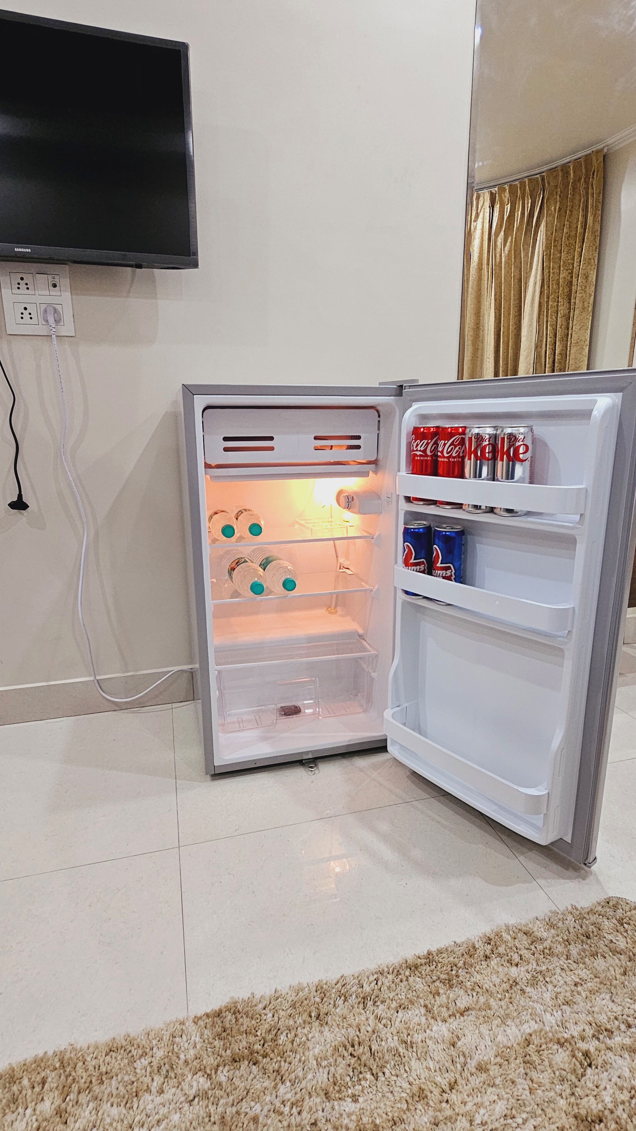 Un refrigerador abierto con bebidas y un televisor montado en la pared.