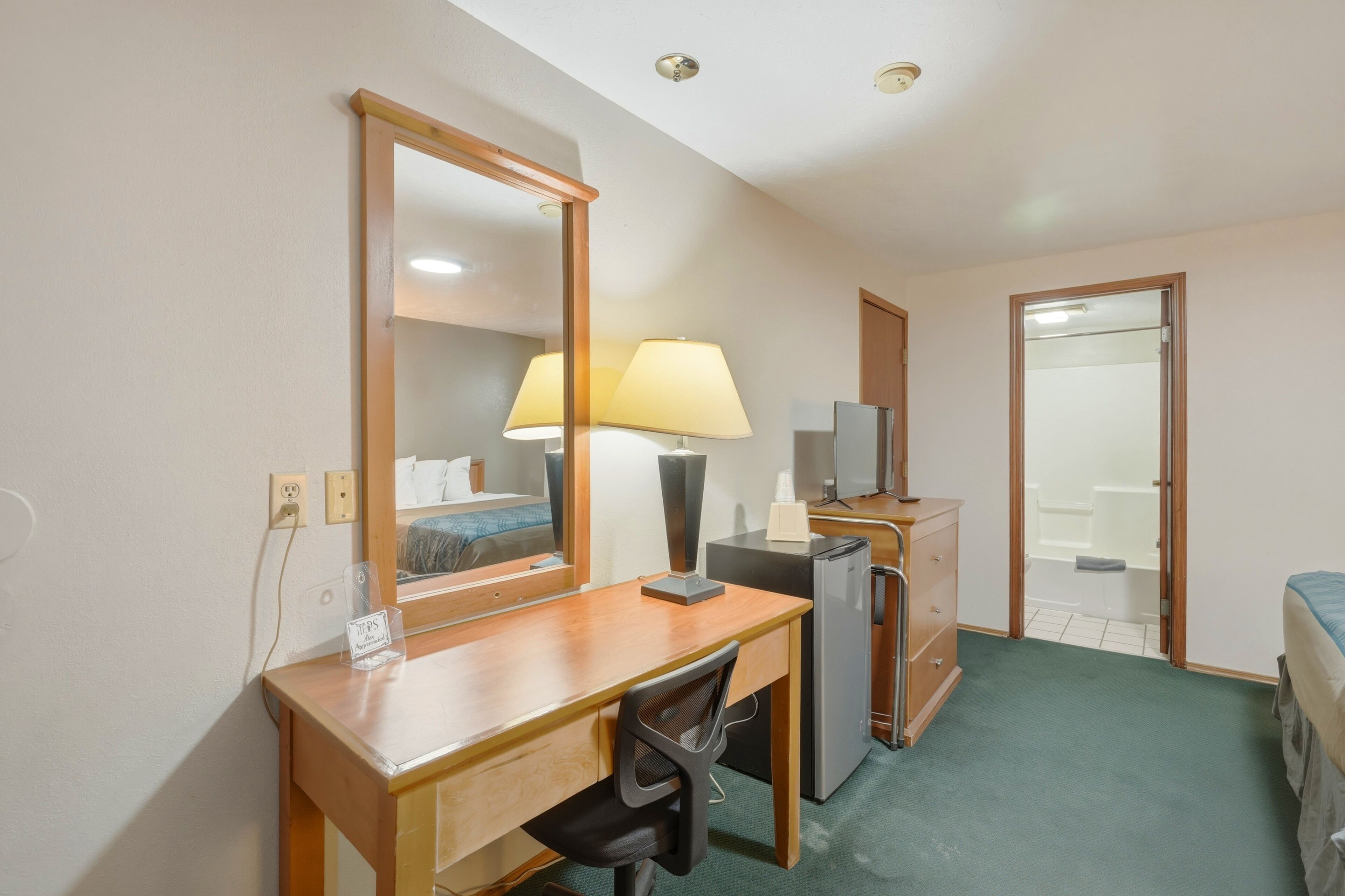 A hotel room with a wooden desk, a chair, a mirror, a lamp, a refrigerator, a dresser, and a bed.