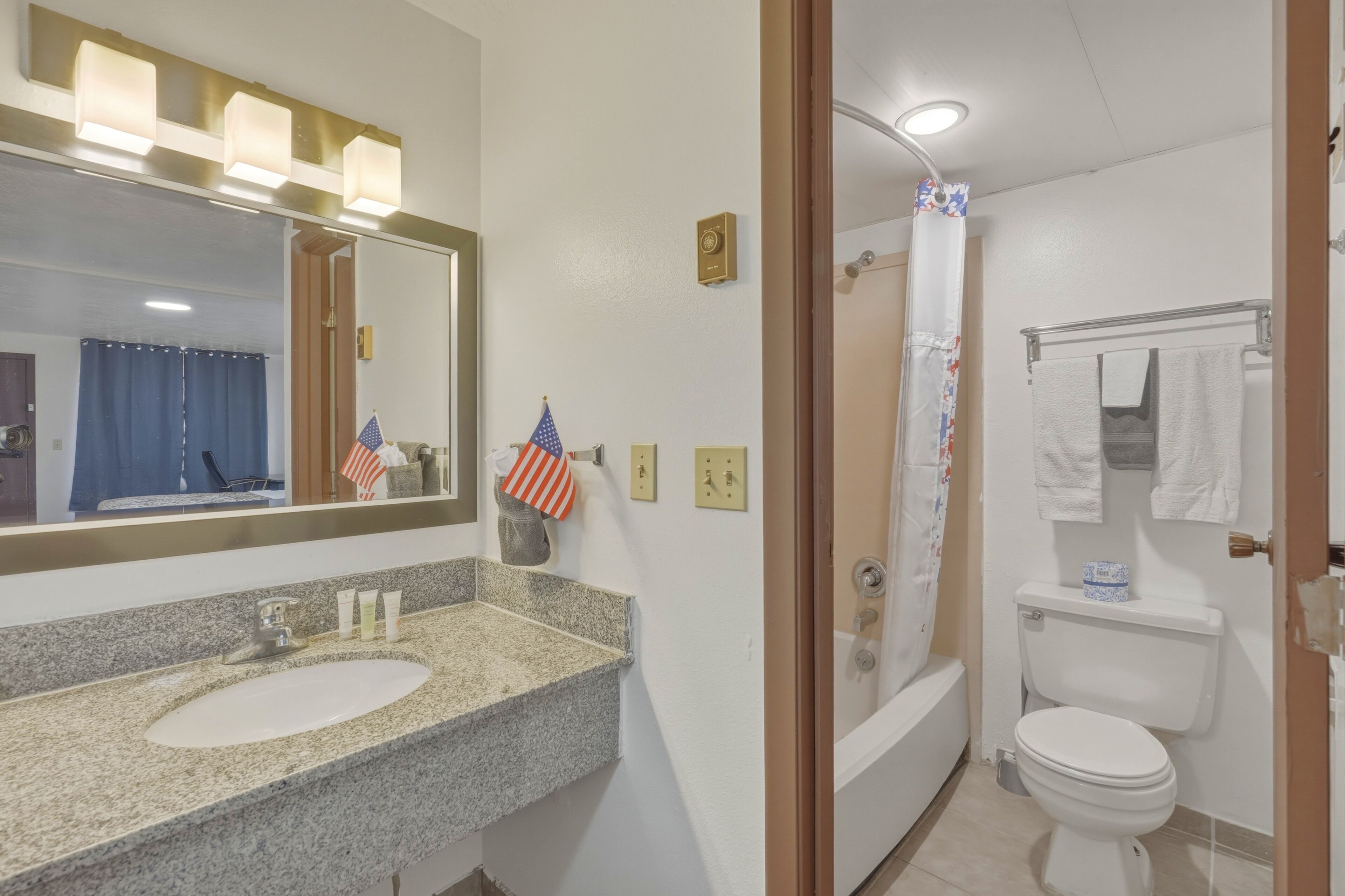 Une salle de bain avec un lavabo doté d’un plan de travail en granit, un miroir, un drapeau américain et un rideau de douche.