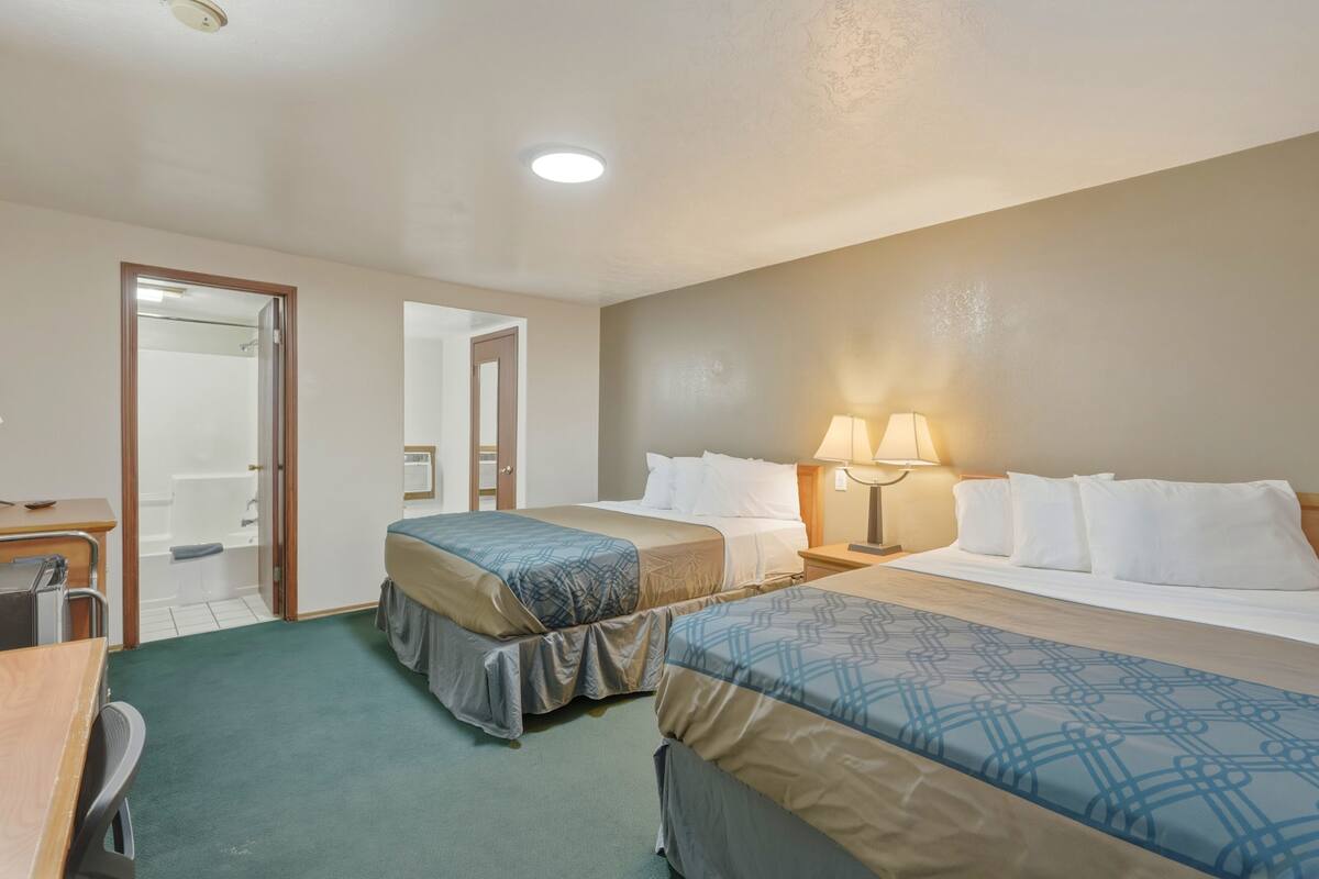 A hotel room with two beds, a desk, a chair, a lamp, and a shower area.
