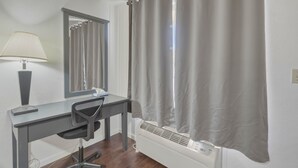 Ett rum med ett skrivbord, en stol, en lampa, en spegel, ett fönster med gardiner och ett element.