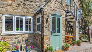 Une petite maison en pierre avec une porte verte, un petit porche agrémenté de plantes en pot et un escalier menant à l’étage supérieur.