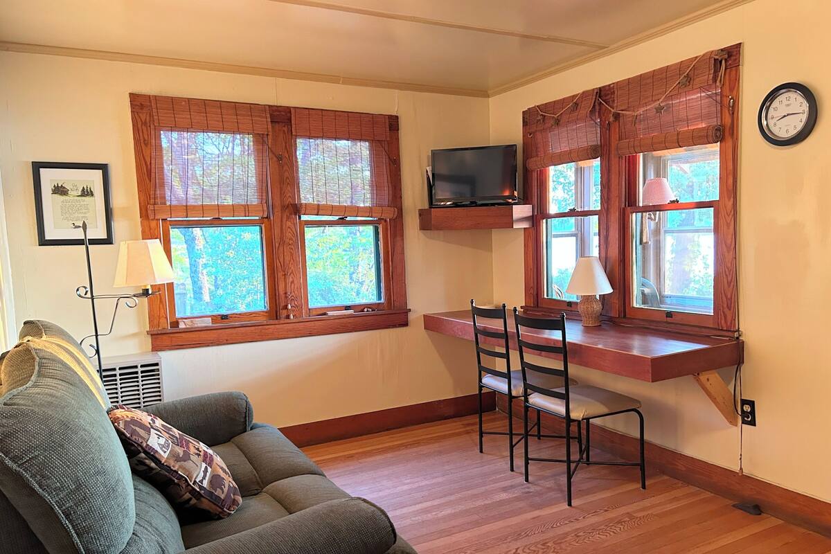 A cozy room with a wooden floor, a sofa with patterned cushions, a dining table with two chairs, and a TV mounted above the table.