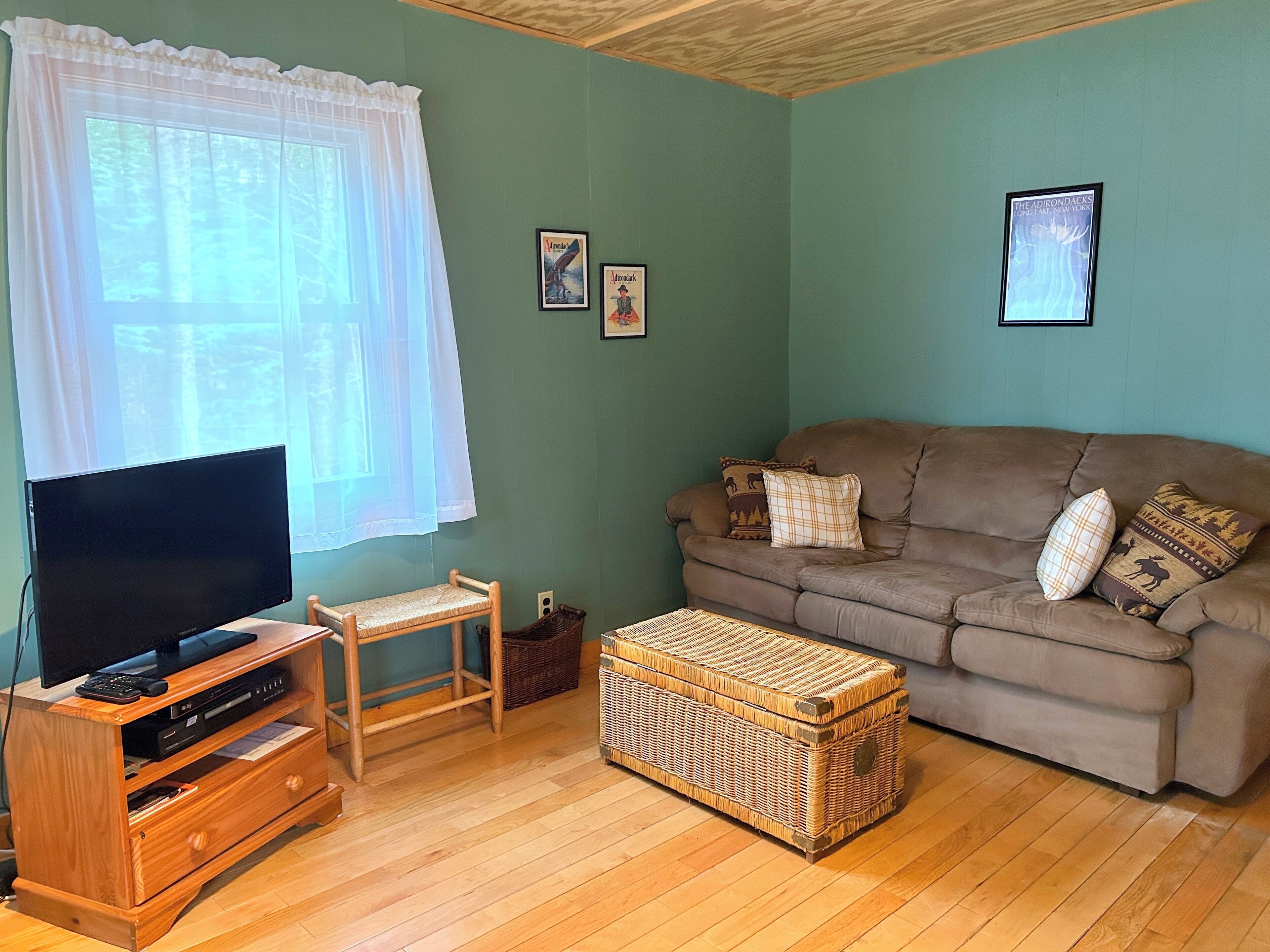 Uma sala de estar com televisão sobre suporte de madeira, um sofá marrom com almofadas estampadas, um banquinho de vime e um pôster emoldurado na parede.