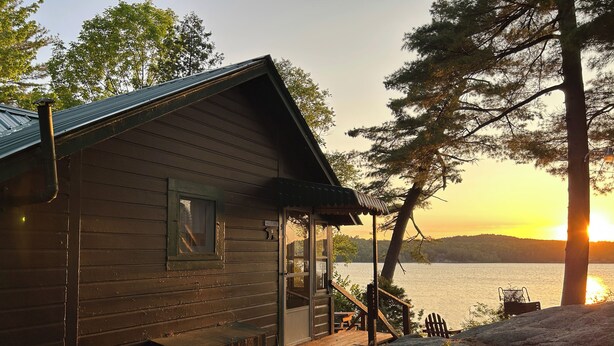 Une cabane avec une véranda, une terrasse en bois et une vue sur un lac au coucher du soleil.