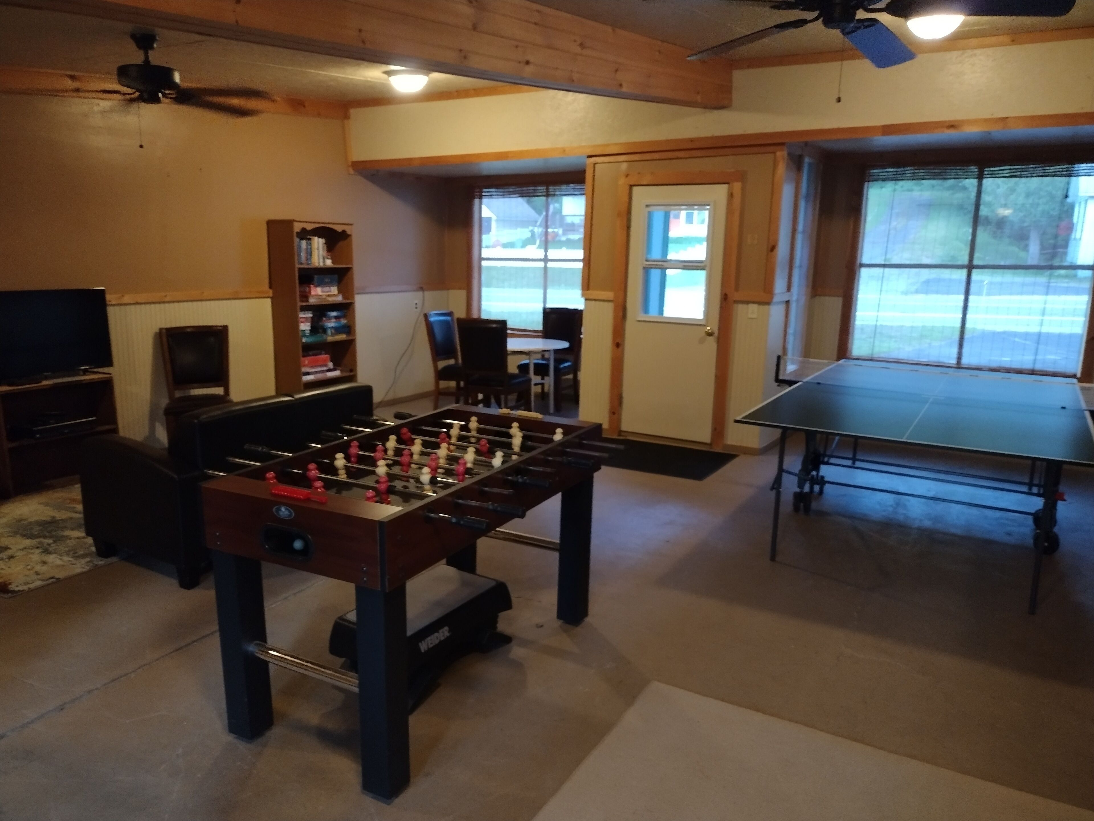 A room with a foosball table, a ping pong table, a television, and a bookshelf.