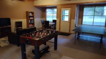 A room with a foosball table, a ping pong table, a television, and a bookshelf.