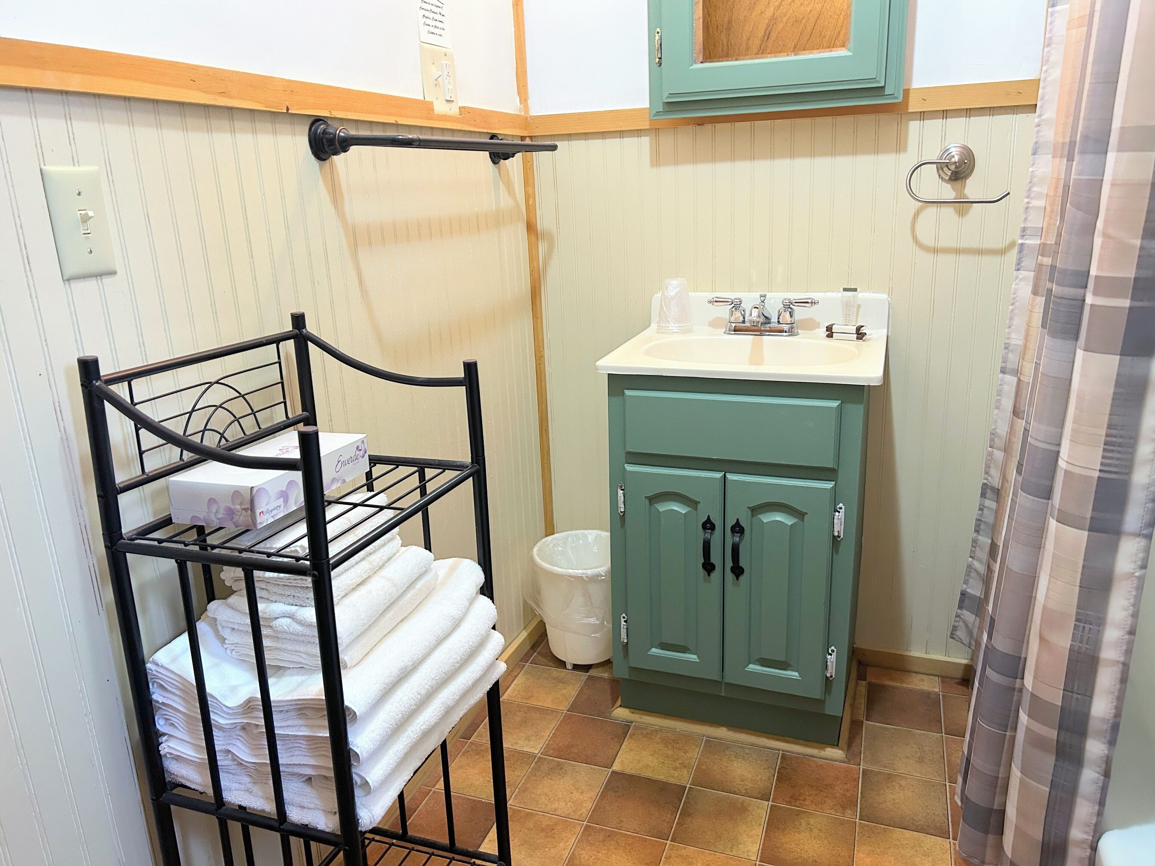 A bathroom with a metal towel rack holding folded towels, a green cabinet with a sink, and a white trash bin.