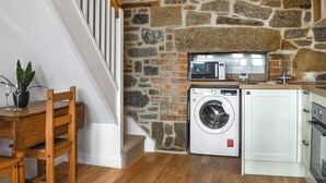 A laundry area with a washing machine and a microwave on a shelf, a wooden chair, and a staircase with white railings. - 1 bedroom accommodation in Penzance (Penzance)