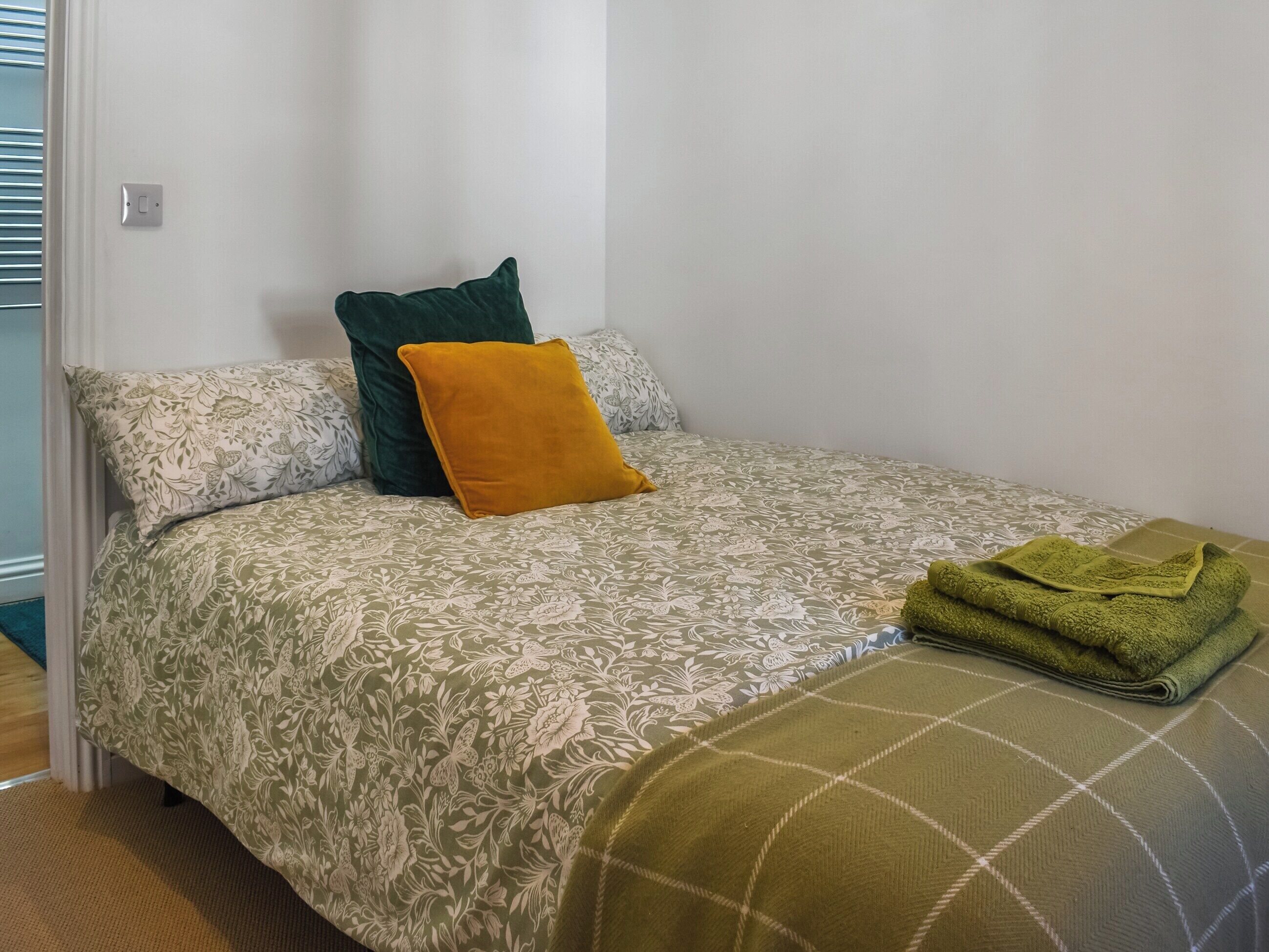 Een eenpersoonsbed met bloemenbedrukte beddengoed, een groen kussen, een mosterdkleurig kussen en een opgevouwen groene handdoek.