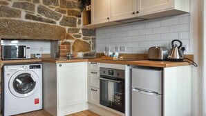 A compact kitchen with a stone wall, white cabinetry, a washing machine, a dishwasher, an oven, and a refrigerator. - 1 bedroom accommodation in Penzance (Penzance)