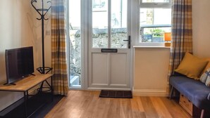 A room with a television on a wooden table, a bench with cushions, a door leading outside, and a window with curtains. - 1 bedroom accommodation in Penzance (Penzance)