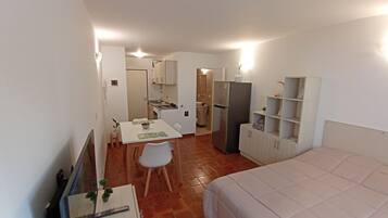Una habitación de hotel compacta con cama, una pequeña mesa de comedor con sillas, kitchenette y baño visible a través de una puerta abierta.