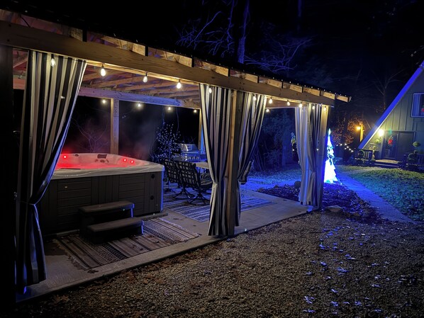 Un jacuzzi bajo una pérgola de madera con luces colgantes, un espacio de descanso cubierto y un árbol de Navidad.