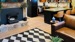 Un interior acogedor con chimenea de piedra, vigas de madera en el techo, una zona de cocina y un espacio de descanso con alfombra a cuadros.