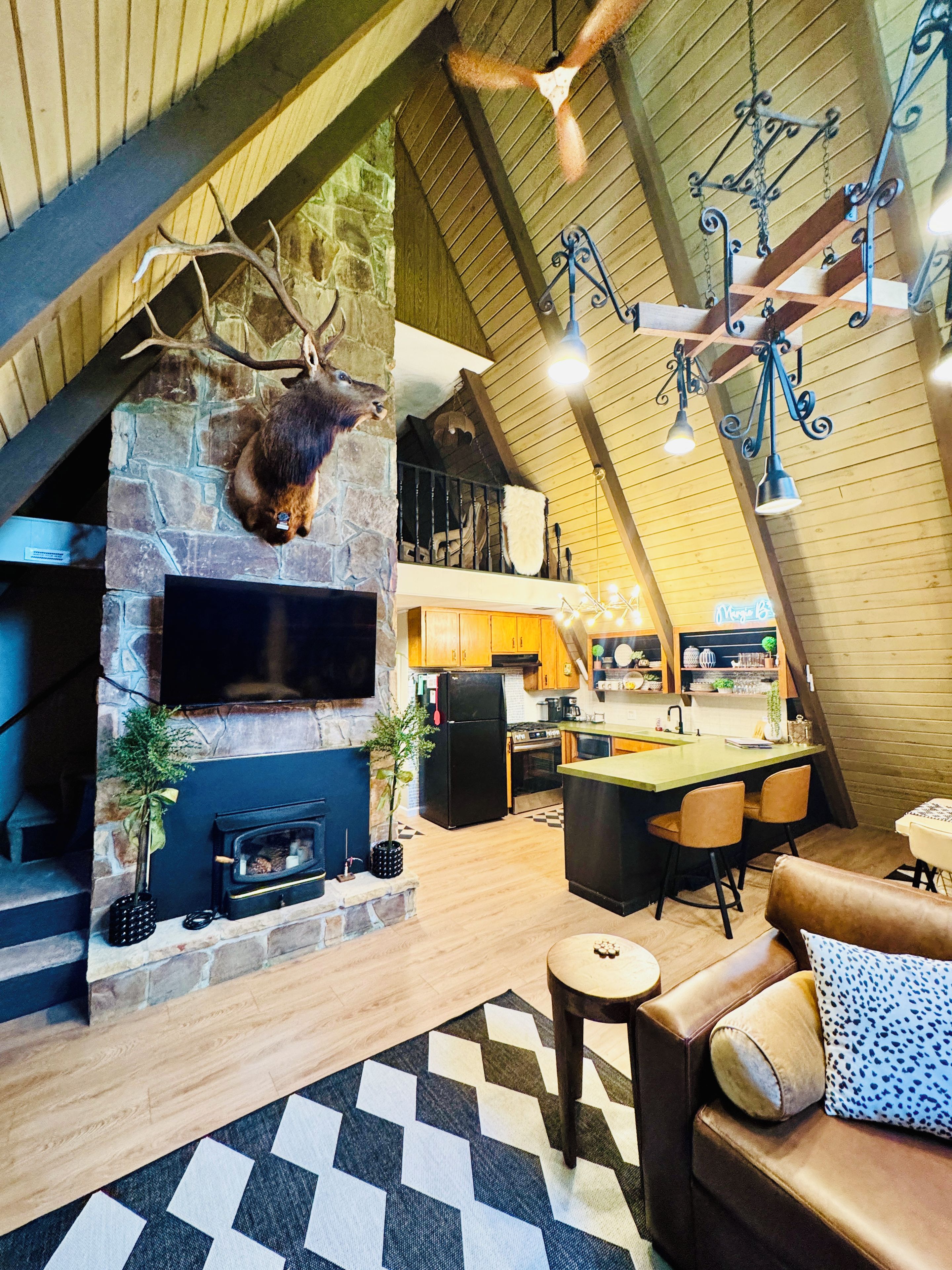 A spacious living area with a stone fireplace, a mounted deer head, a flat-screen TV, a dining area with a bar, and a loft with a sloped ceiling.