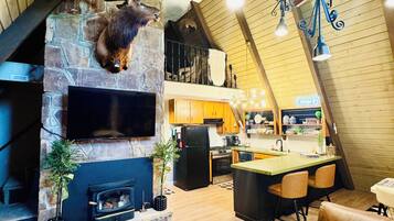 A spacious living area with a stone fireplace, a mounted deer head, a flat-screen TV, a dining area with a bar, and a loft with a sloped ceiling.