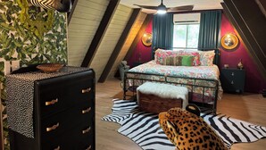 Ein Schlafzimmer mit schräg stehender Decke, einem zebraartig gemusterten Teppich, einem leopardenmuster Plüschtier, einem Bett mit farbenfroher Bettwäsche, einem Schminktisch und einem an der Wand befestigten Spiegel.