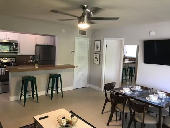 A modern kitchen with a central island, bar stools, a dining area with a table set for four, and a ceiling fan.
