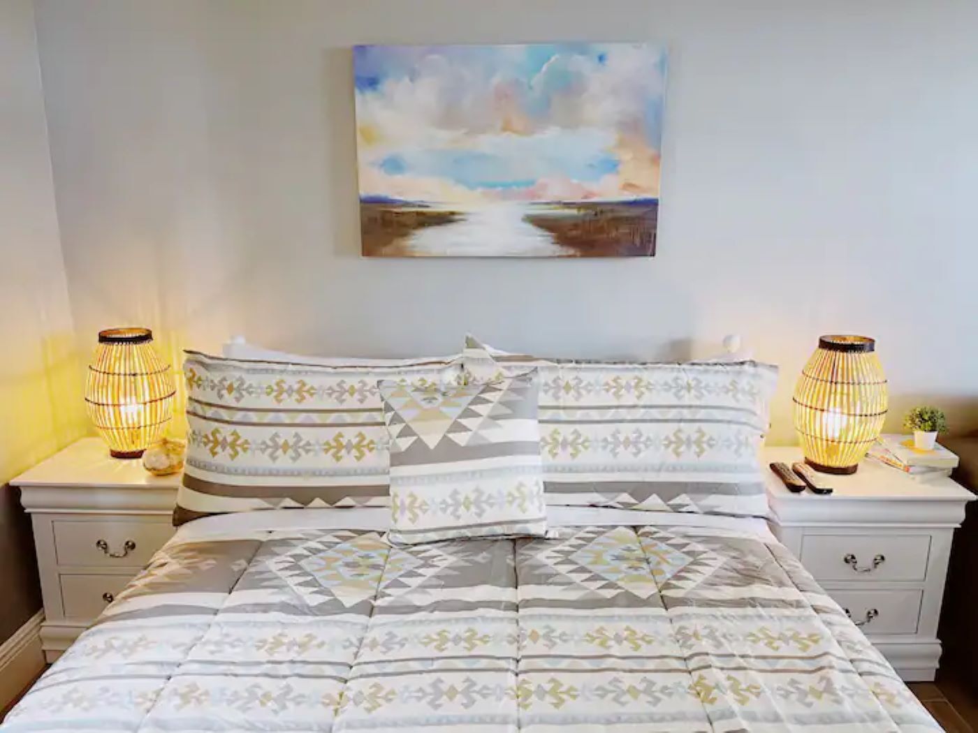 A neatly made bed with patterned bedding, two bedside lamps, and a painting above the bed.