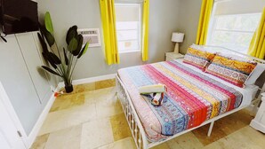 A bedroom with a bed, a nightstand, a lamp, a potted plant, and a window with yellow curtains. - Cozy 1 Bedroom APT 1 block off the beach W/ pool (pompano beach)