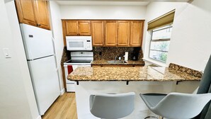 A compact kitchen with wooden cabinets, a white refrigerator, a microwave, a stove, and a granite countertop. There are two bar stools in front of the counter. - Trendy, Budget-Friendly 1 Bedroom Apt, Pool Access (Fort Lauderdale)