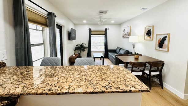 A modern kitchen with granite countertops, a dining area with a wooden table and chairs, a sofa, and a TV mounted on the wall. - Trendy, Budget-Friendly 1 Bedroom Apt, Pool Access (Fort Lauderdale)