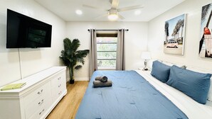 A bedroom with a bed, a TV, a ceiling fan, a window with curtains, a plant, and a wall with pictures. - Trendy, Budget-Friendly 1 Bedroom Apt, Pool Access (Fort Lauderdale)