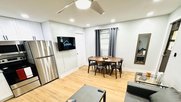 A modern kitchen with stainless steel appliances, a dining area with a wooden table and chairs, a grey sofa, and a flat-screen TV mounted on the wall. - Trendy & Central 2 Bedroom Apt (Fort Lauderdale)