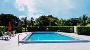 A rectangular swimming pool with a ladder, surrounded by a white sandy area. There are chairs and a red umbrella near the pool.
