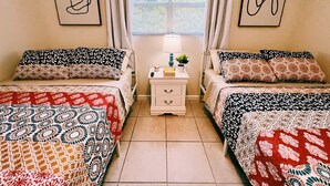 Une chambre à coucher avec deux lits, chacun agrémenté de draps à motifs, une table de chevet avec une lampe et une plante, et des œuvres d’art accrochées aux murs.