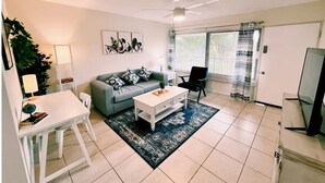 A living room with a sofa, coffee table, and a TV. There is a ceiling fan and a window with curtains.