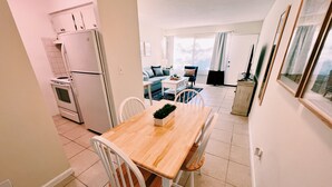 Uma cozinha compacta com eletrodomésticos brancos, uma área de jantar com mesa de madeira e cadeiras brancas, e um espaço de estar com sofá e televisão.