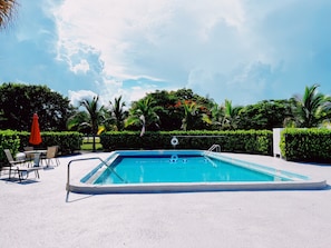 Una piscina rectangular con una escalera, rodeada por una zona de arena blanca. Hay sillas y una sombrilla roja cerca de la piscina.