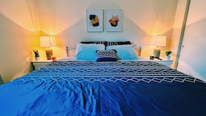 Uma cama bem arrumada com colcha azul estampada, dois quadros pendurados na parede e abajures de cada lado.