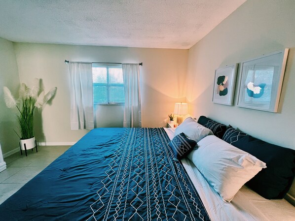 A bedroom with a large bed, a bedside table with a lamp, a potted plant, and a window with curtains.