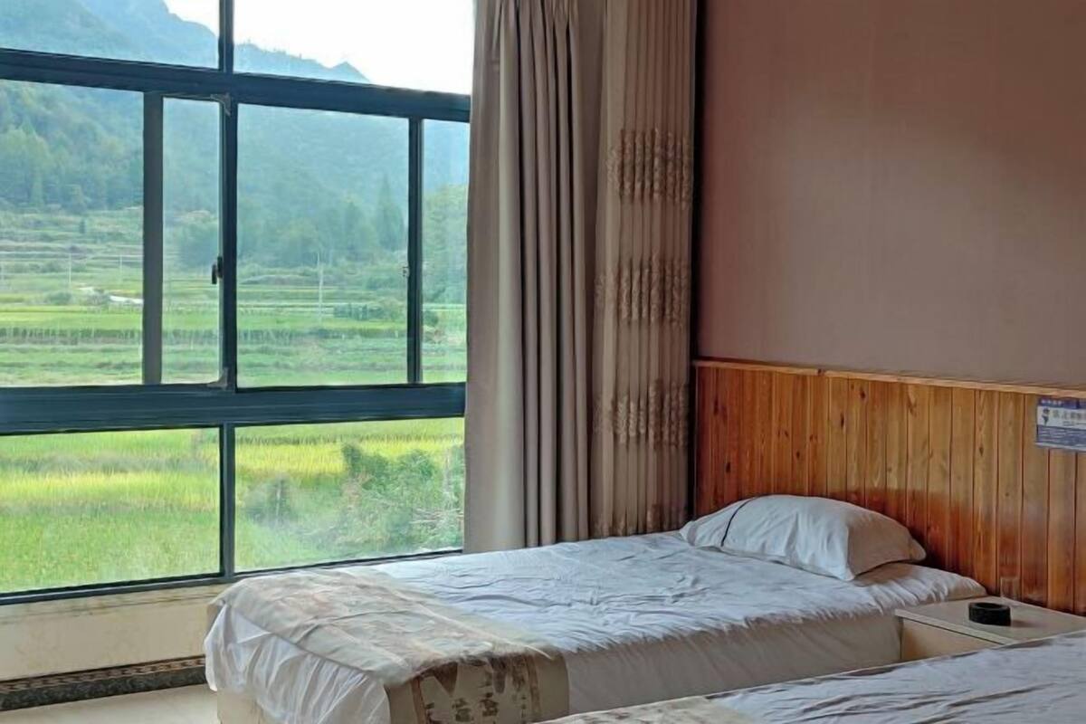 Una habitación con un ventanal grande que ofrece vistas a un paisaje verde, una cama con cabecera de madera y una mesita.