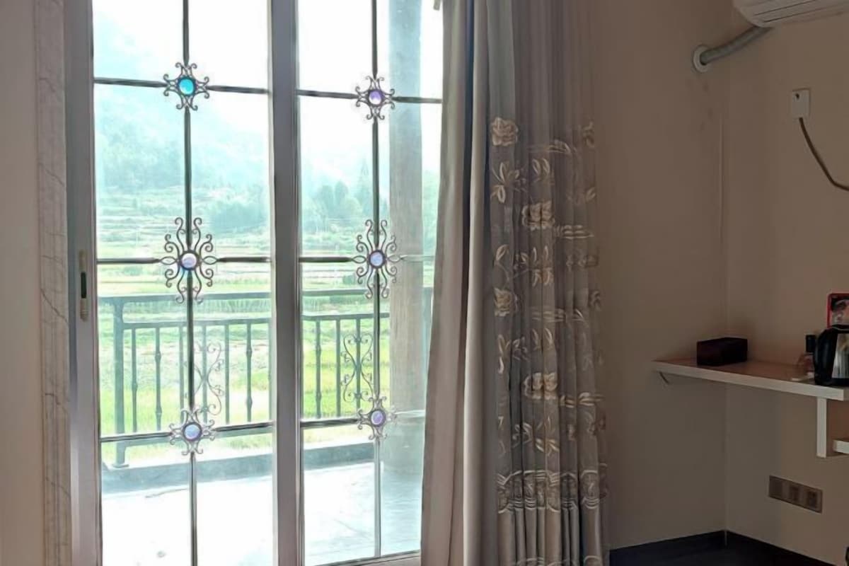 Habitación con un ventanal grande que presenta celosías metálicas decorativas y vistas a un paisaje verde. Se observa una repisa blanca con una tetera y una caja.