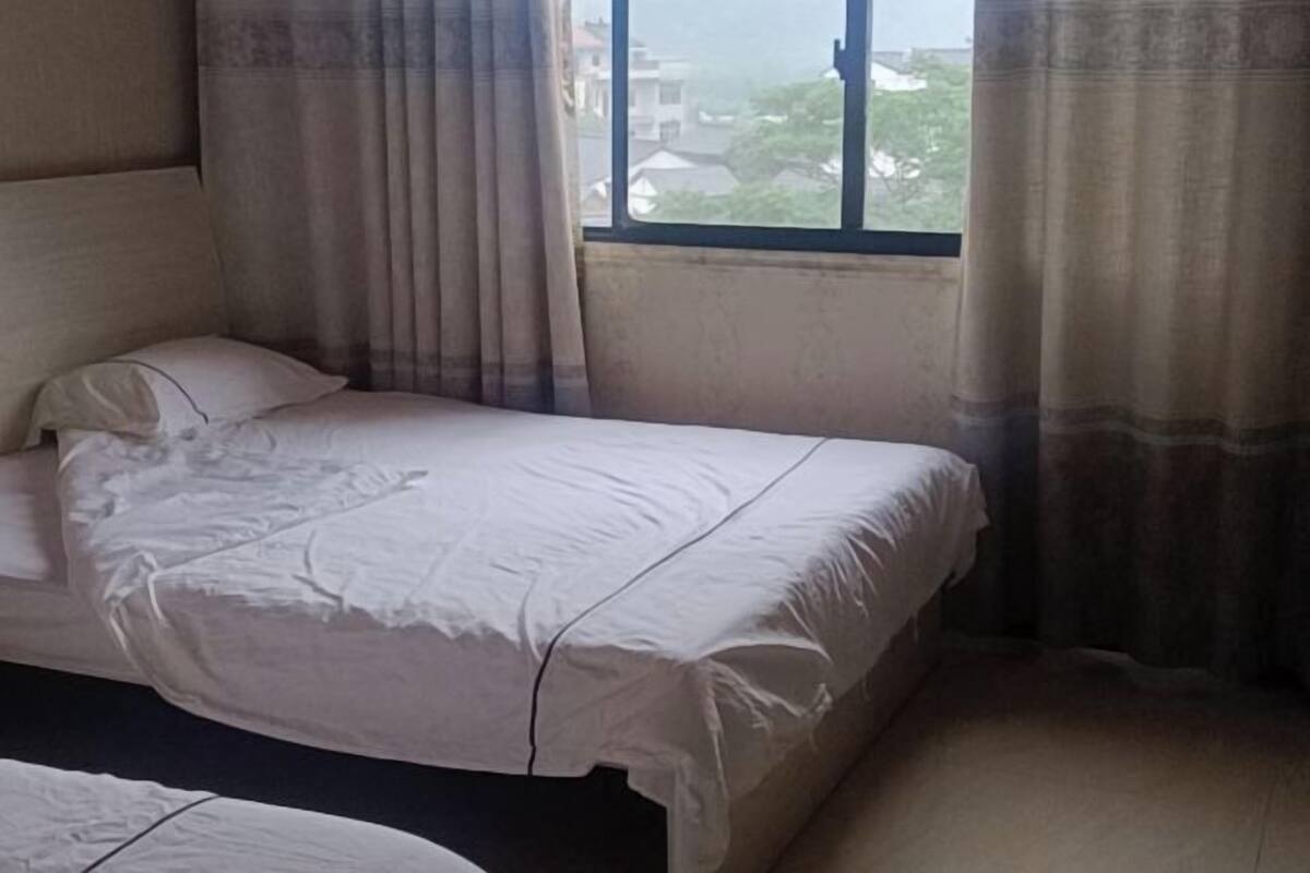 Una cama individual con una colcha blanca en una habitación con ventana y cortinas.