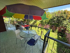 Ein Balkon mit rundem Tisch und Stühlen, einem bunten Sonnenschirm und Blick auf Grünflächen sowie ein rotes Fahrzeug im Hintergrund.