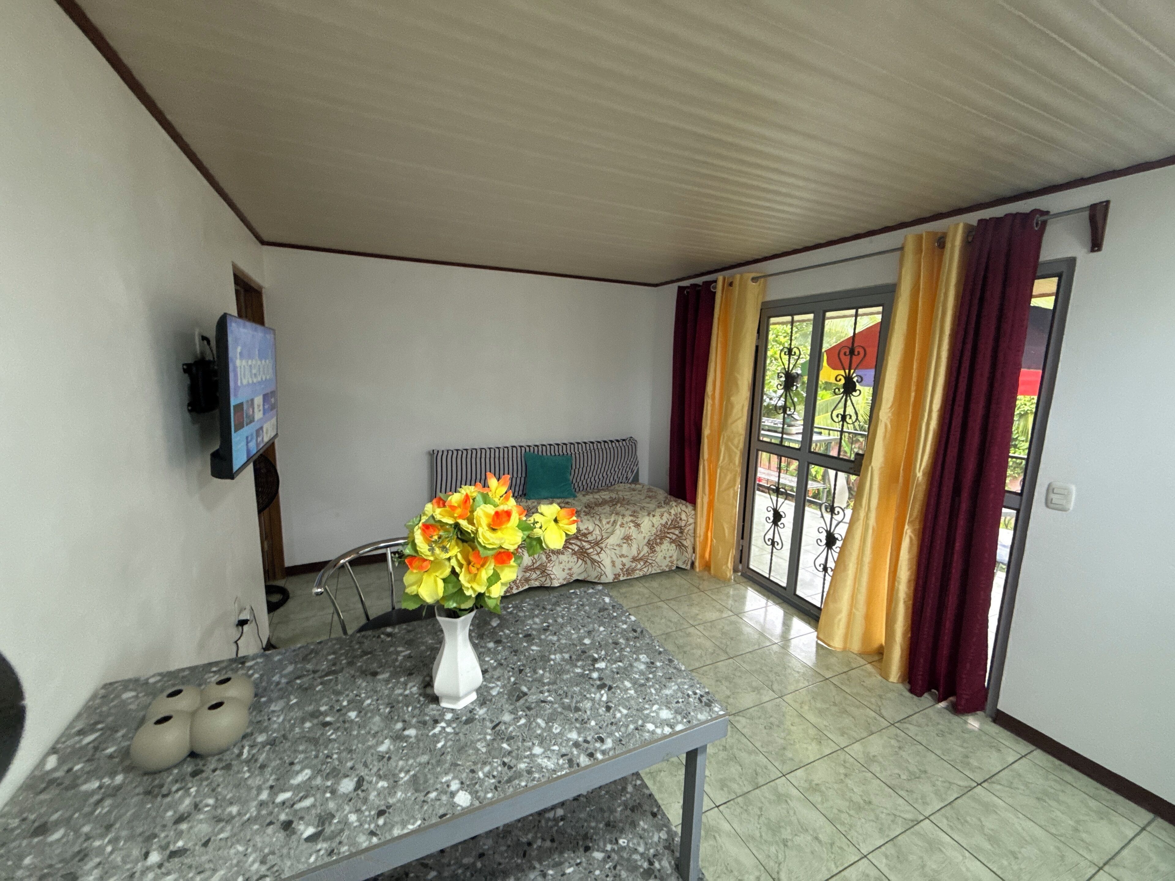 Ein Zimmer mit einem Bett, einem Tisch mit Blumen in einer Vase, einem Fernseher an der Wand und einer Glastür mit Vorhängen.