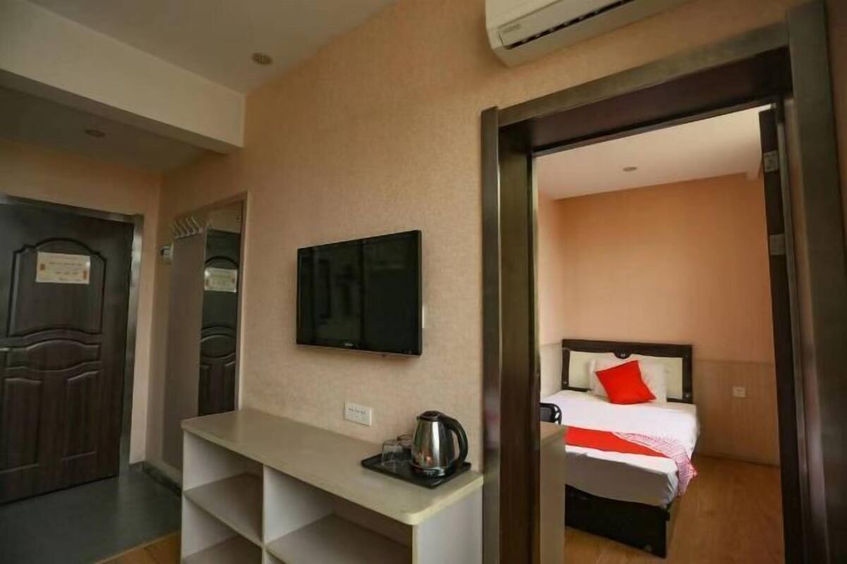 A hotel room with a bed, a TV mounted on the wall, a desk with a kettle, and a door leading to another room.