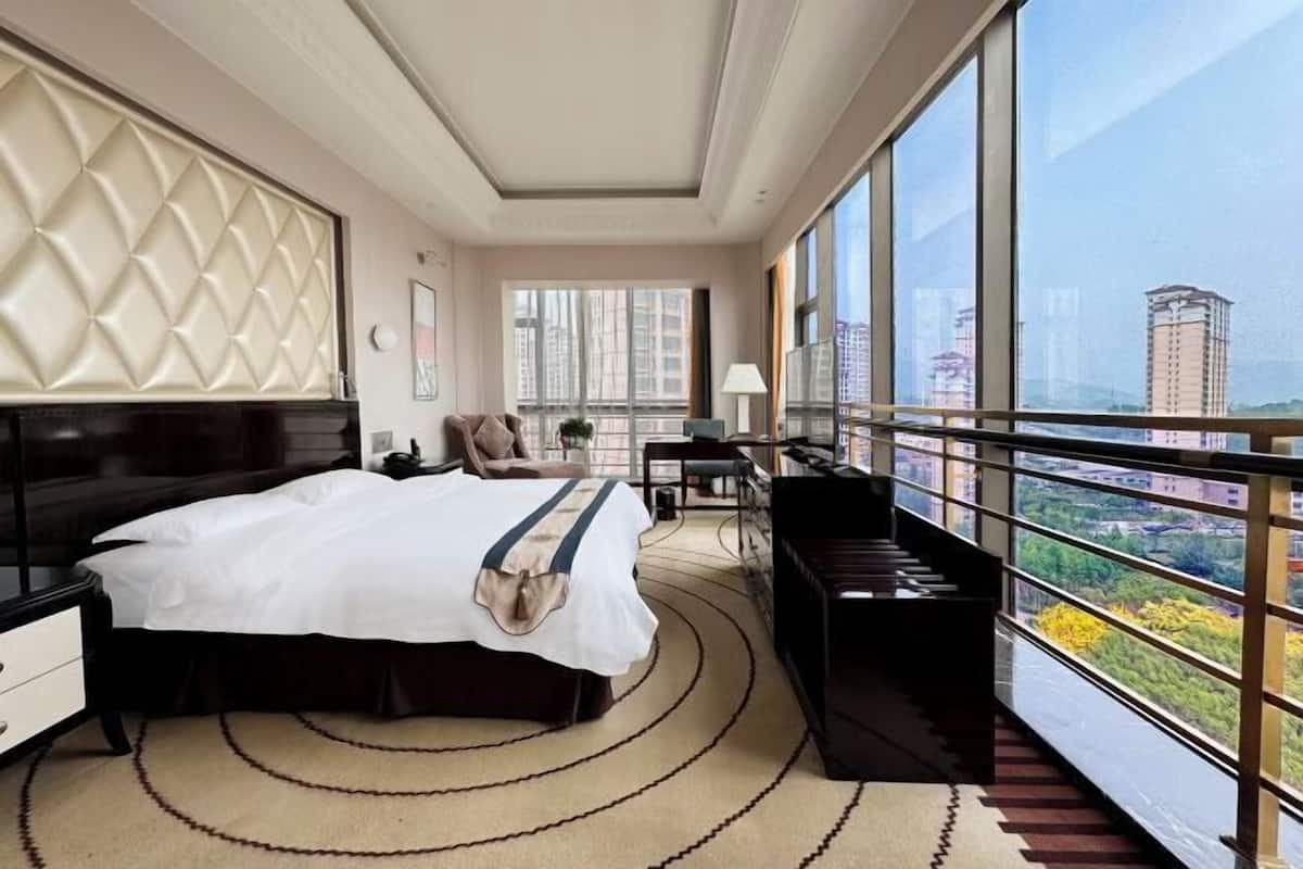 Une chambre d’hôtel avec un grand lit, un piano, un bureau et une vue sur la ville grâce à de grandes fenêtres.
