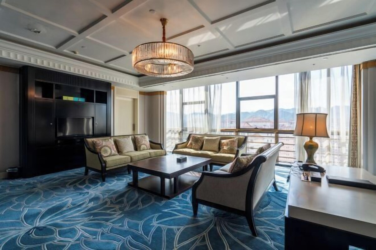 Amplia habitación de hotel con un gran ventanal con vistas a la montaña, un candelabro, un conjunto de sofás y un televisor de pantalla plana.