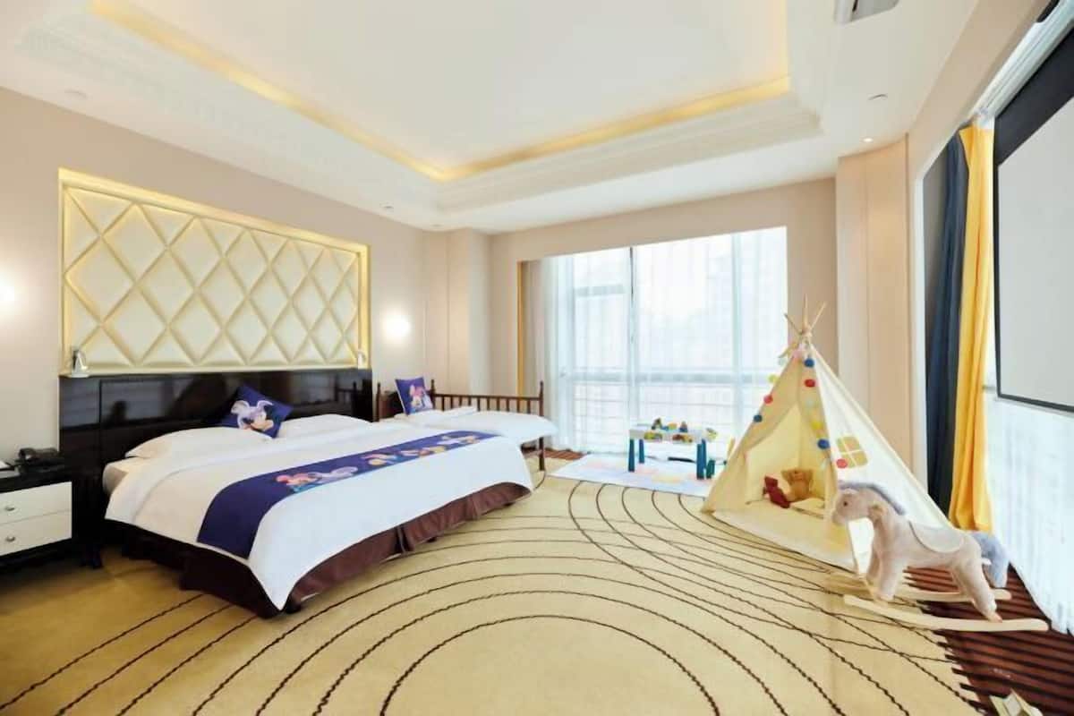 Une chambre d’hôtel avec un grand lit, une tête de lit ornée d’un motif en losanges, une fenêtre avec des rideaux, un petit espace de jeux avec un tipi et des jouets, et un plancher en bois.
