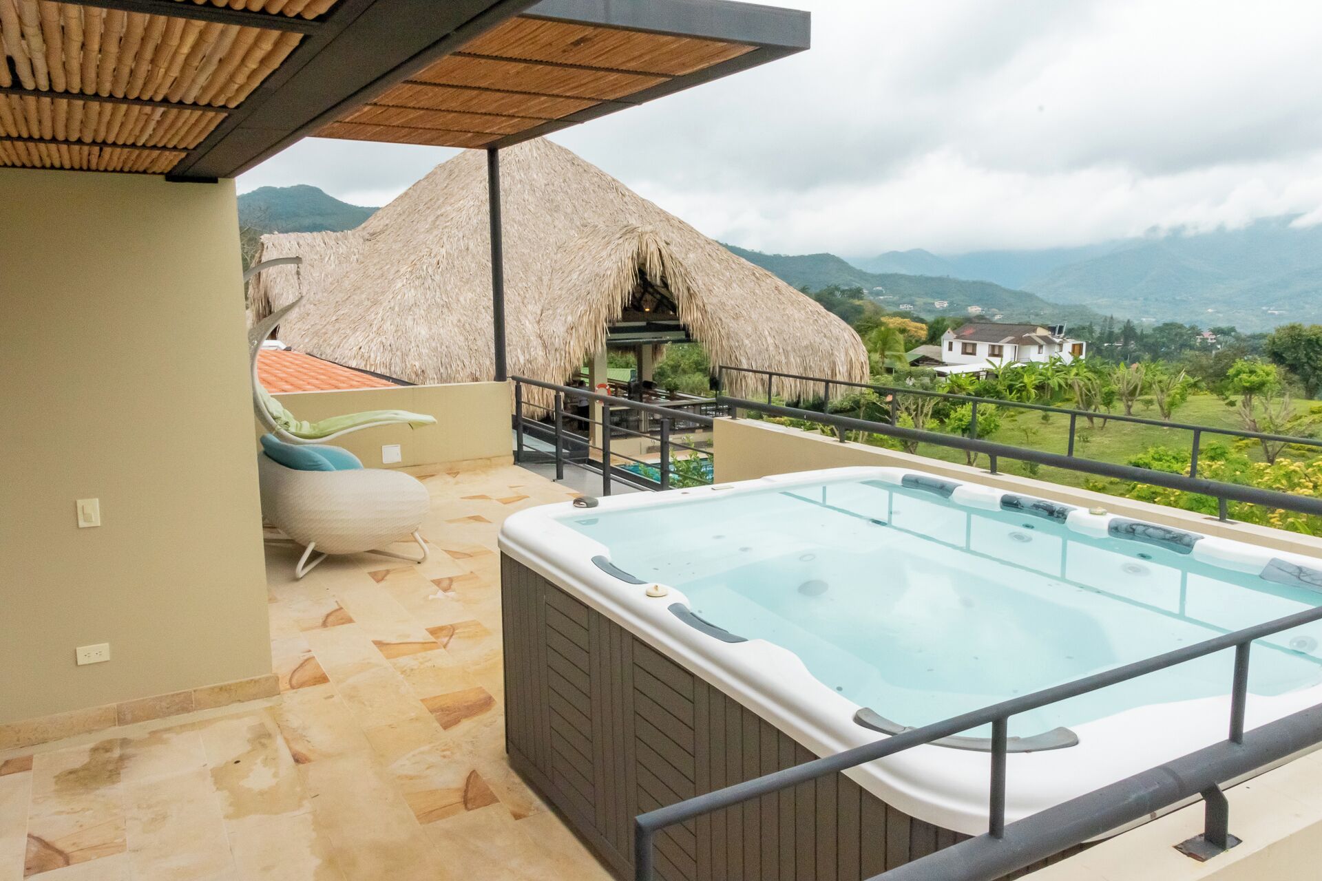 Un balcón con un jacuzzi, una estructura con techo de paja y vistas a las montañas.