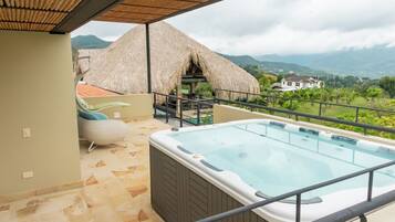 Un balcón con un jacuzzi, una estructura con techo de paja y vistas a las montañas.