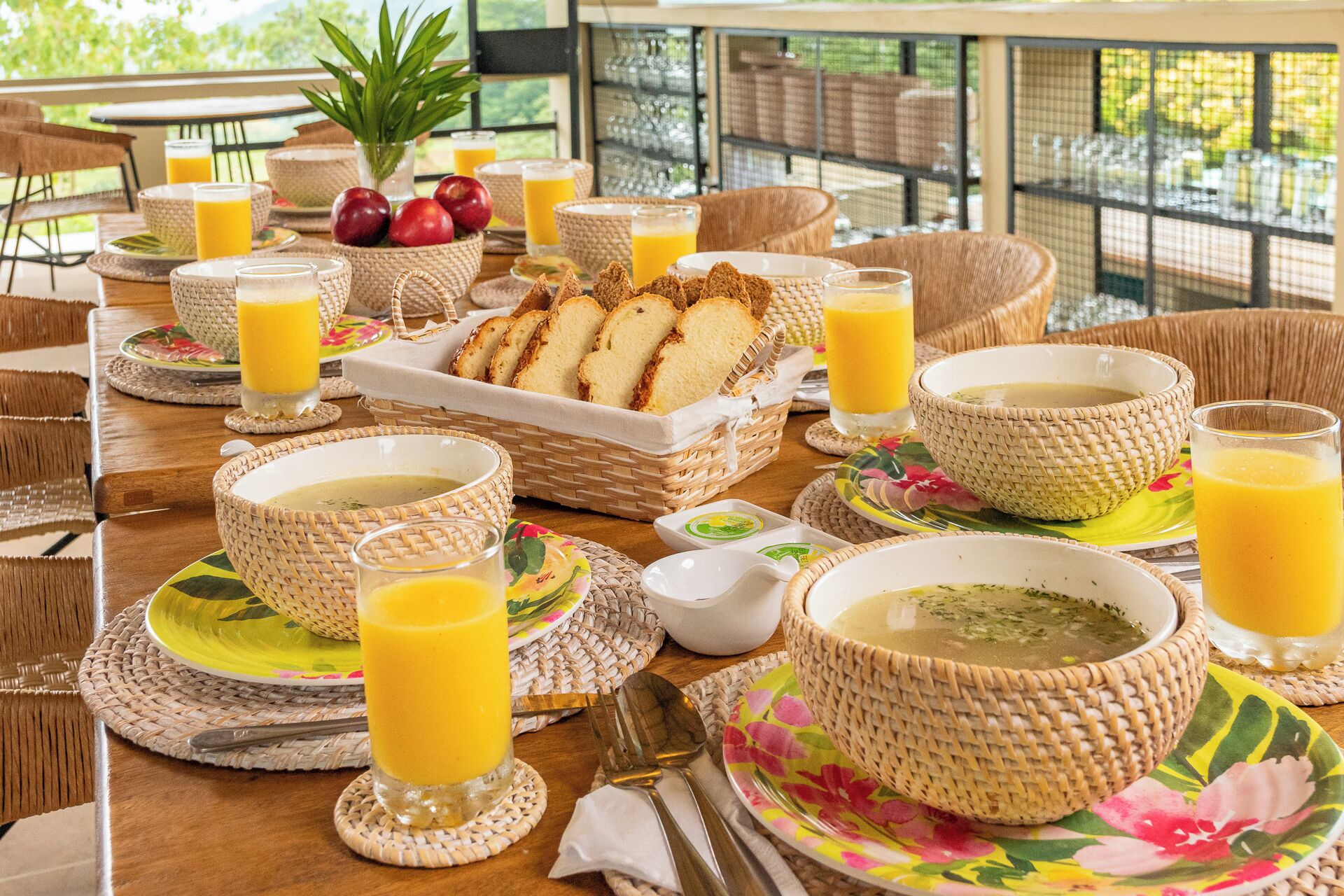 Un desayuno con cesteras de mimbre, tazones y vasos de jugo de naranja sobre una mesa de madera.